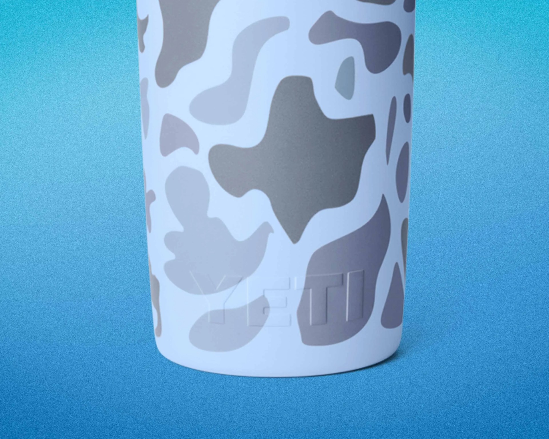 Bottom portion of a white YETI tumbler with gray camouflage pattern on a blue gradient background.