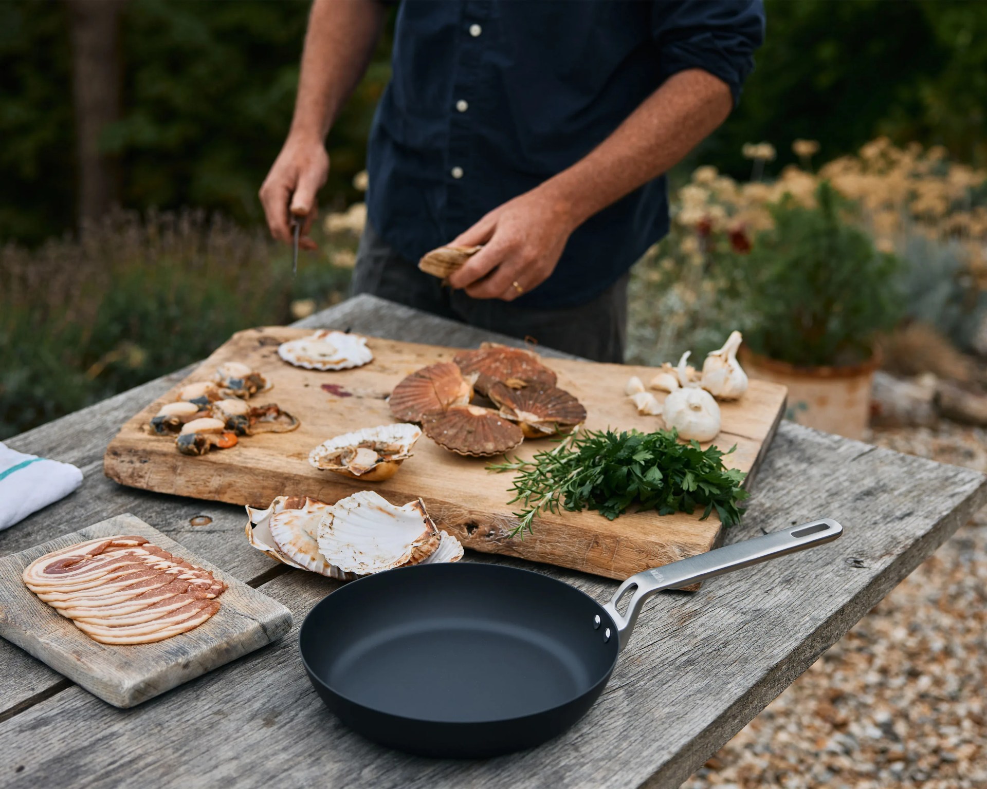 Black frying pan with metal handle on a wooden table next to sliced bacon, scallop shells, herbs, and garlic.