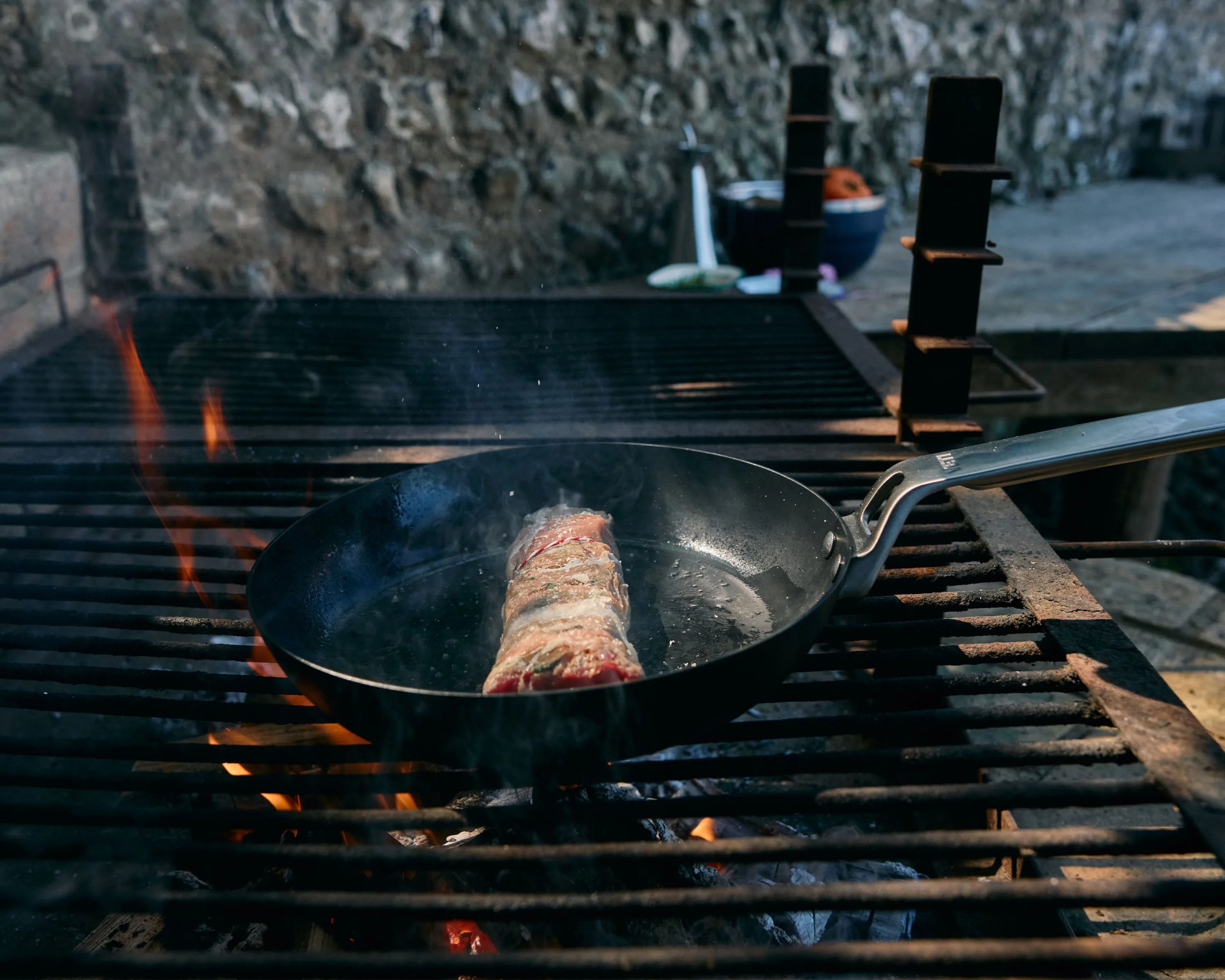Black frying pan with a seasoned meat roll cooking over an open flame grill outdoors.