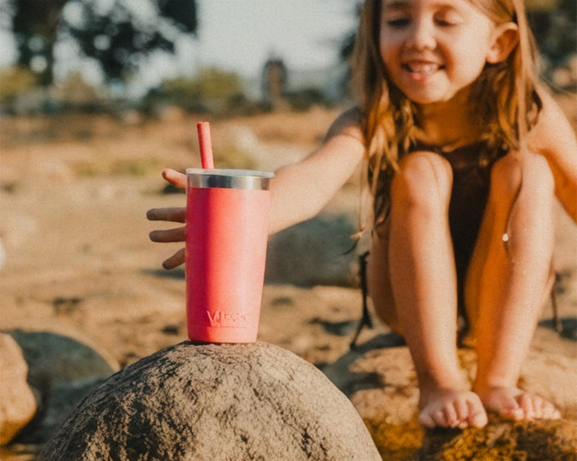 Pink insulated tumbler with a metal lid and straw on a rock near a seated child outdoors.