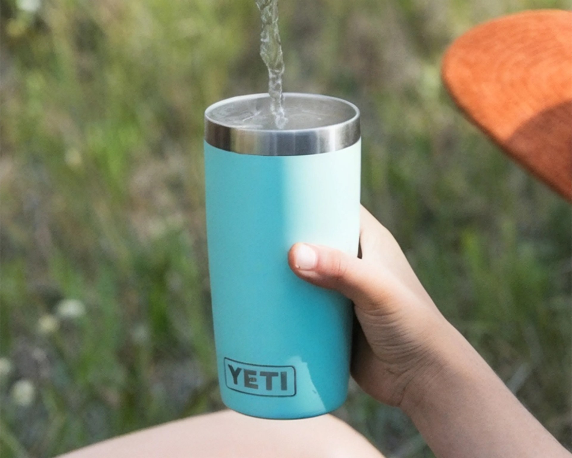 Light blue YETI tumbler with stainless steel rim held in a hand outdoors with water being poured into it.