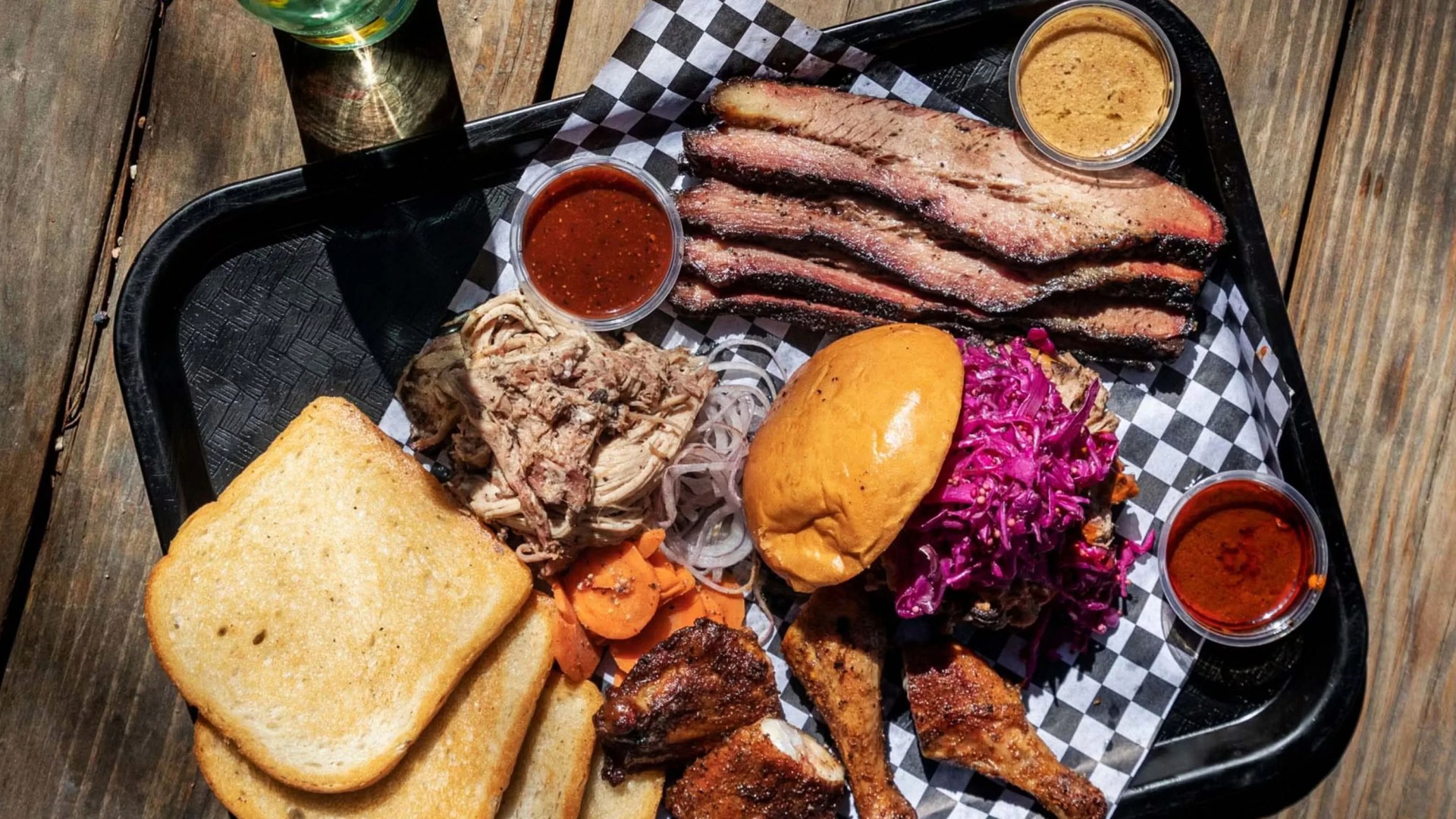 Black tray with sliced smoked brisket, pulled pork, toasted bread, chicken pieces, sandwich with purple slaw, pickled carrots, onions, and four small containers of barbecue sauces.
