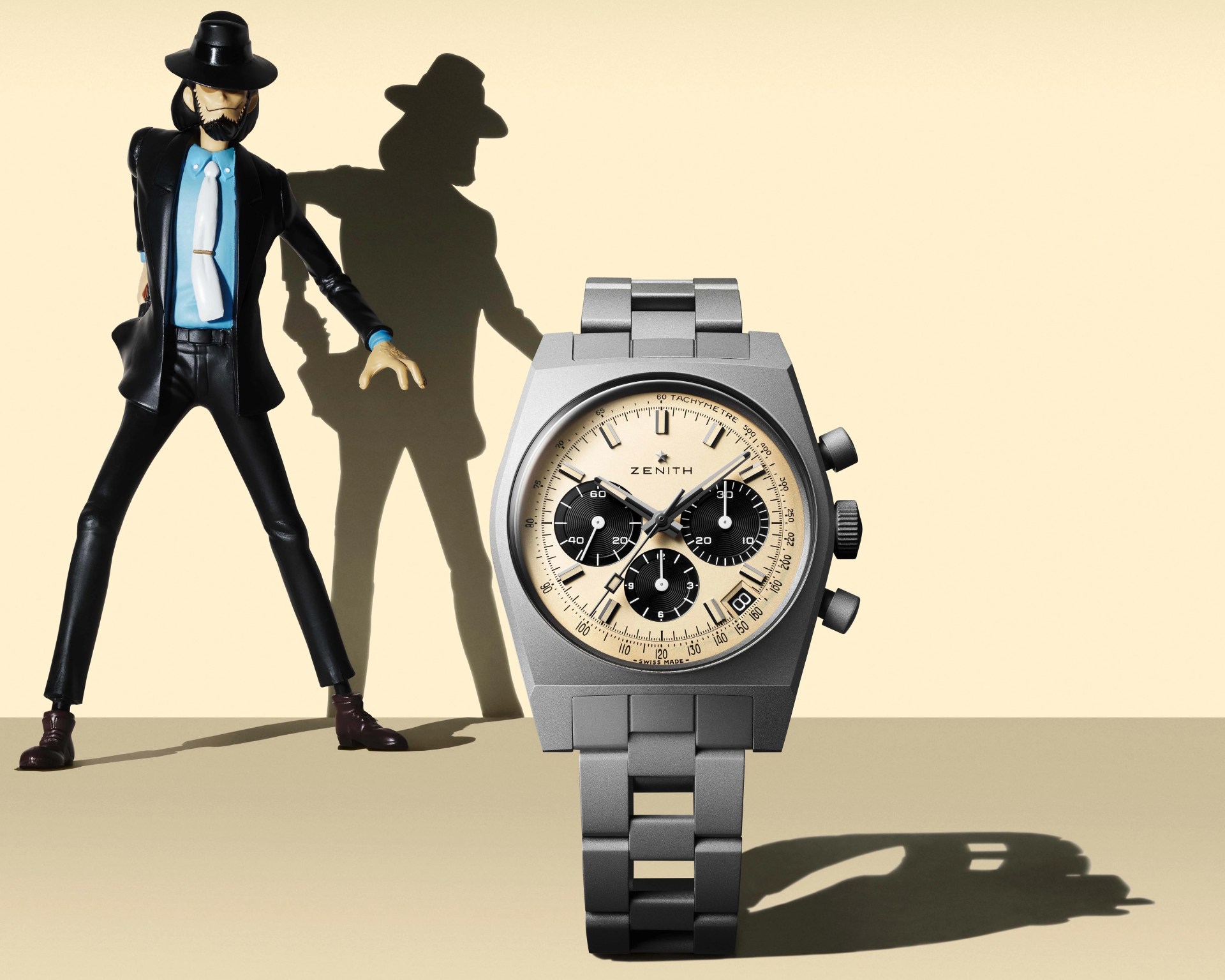 Matte silver Zenith chronograph watch with beige dial and black subdials next to a stylized figure in black suit and hat.