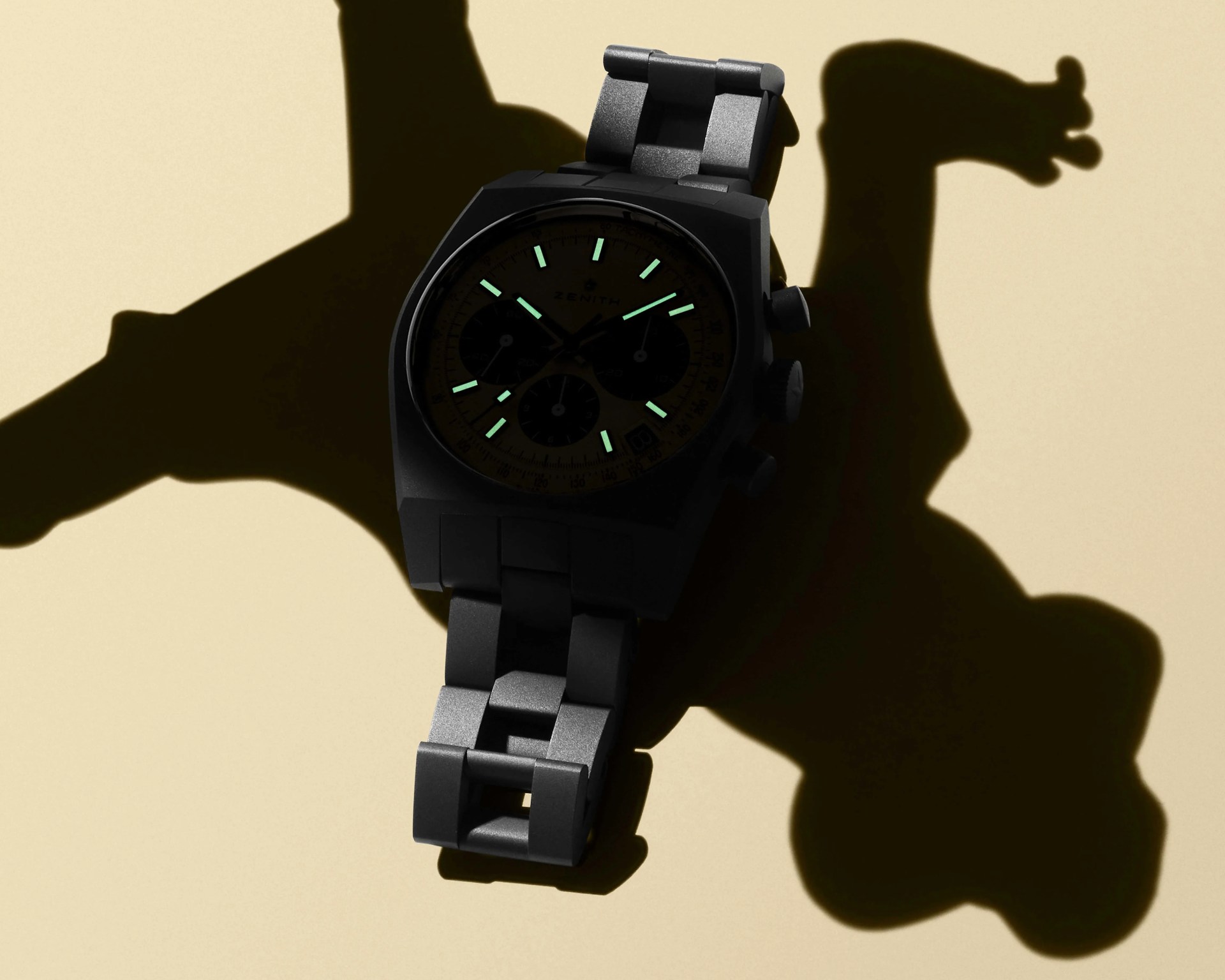 Black metal wristwatch with glowing green hour markers and hands casting a shadow on a beige surface.