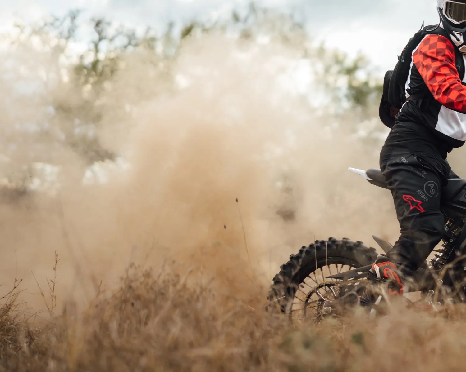 Dirt bike rear wheel kicking up dust with rider wearing black and red gear in dry grass.