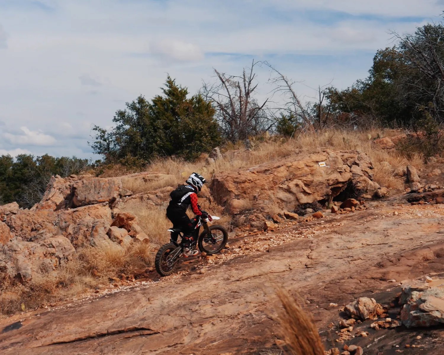 Person wearing a black and red outfit riding a dirt bike uphill on a rocky trail with dry grass and trees.