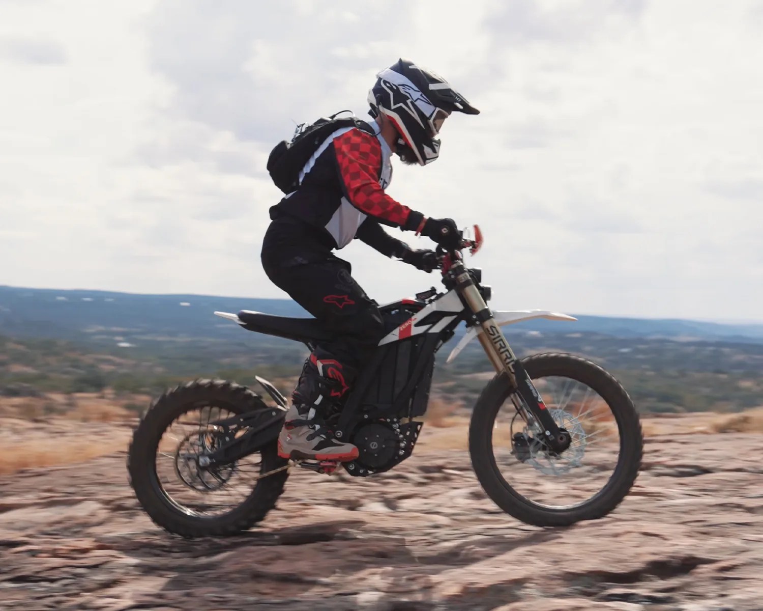 Person wearing black and red motocross gear riding a white and black off-road electric dirt bike on rocky terrain.
