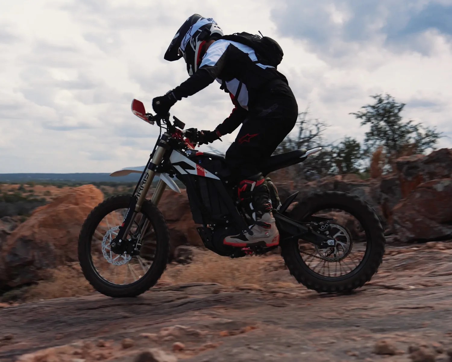 Person wearing black and white gear riding a black and white off-road electric dirt bike on rocky terrain.