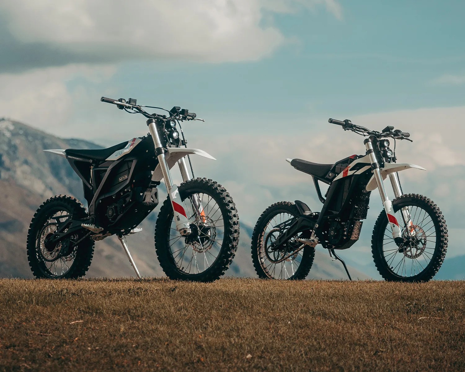Two black and white electric dirt bikes with knobby tires parked on grass with mountains and cloudy sky in the background.