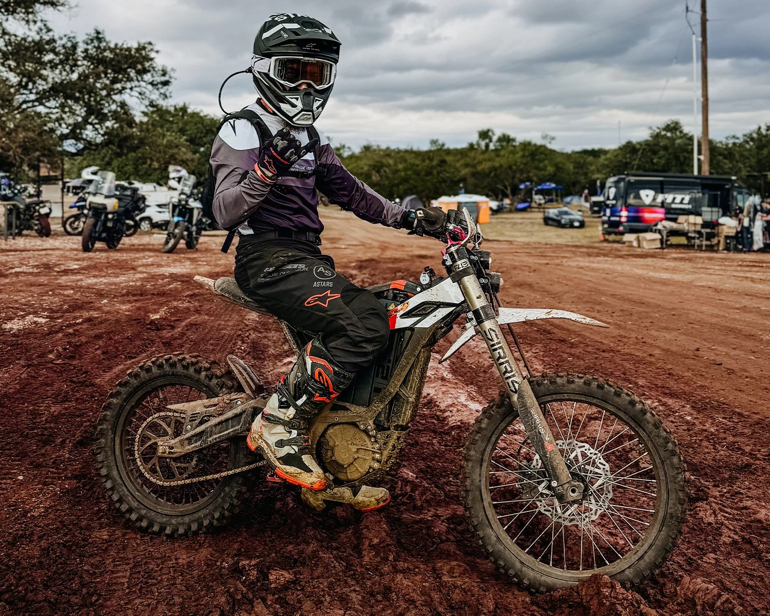 Person wearing black and white motocross gear and helmet sitting on a muddy off-road electric dirt bike on a dirt track.