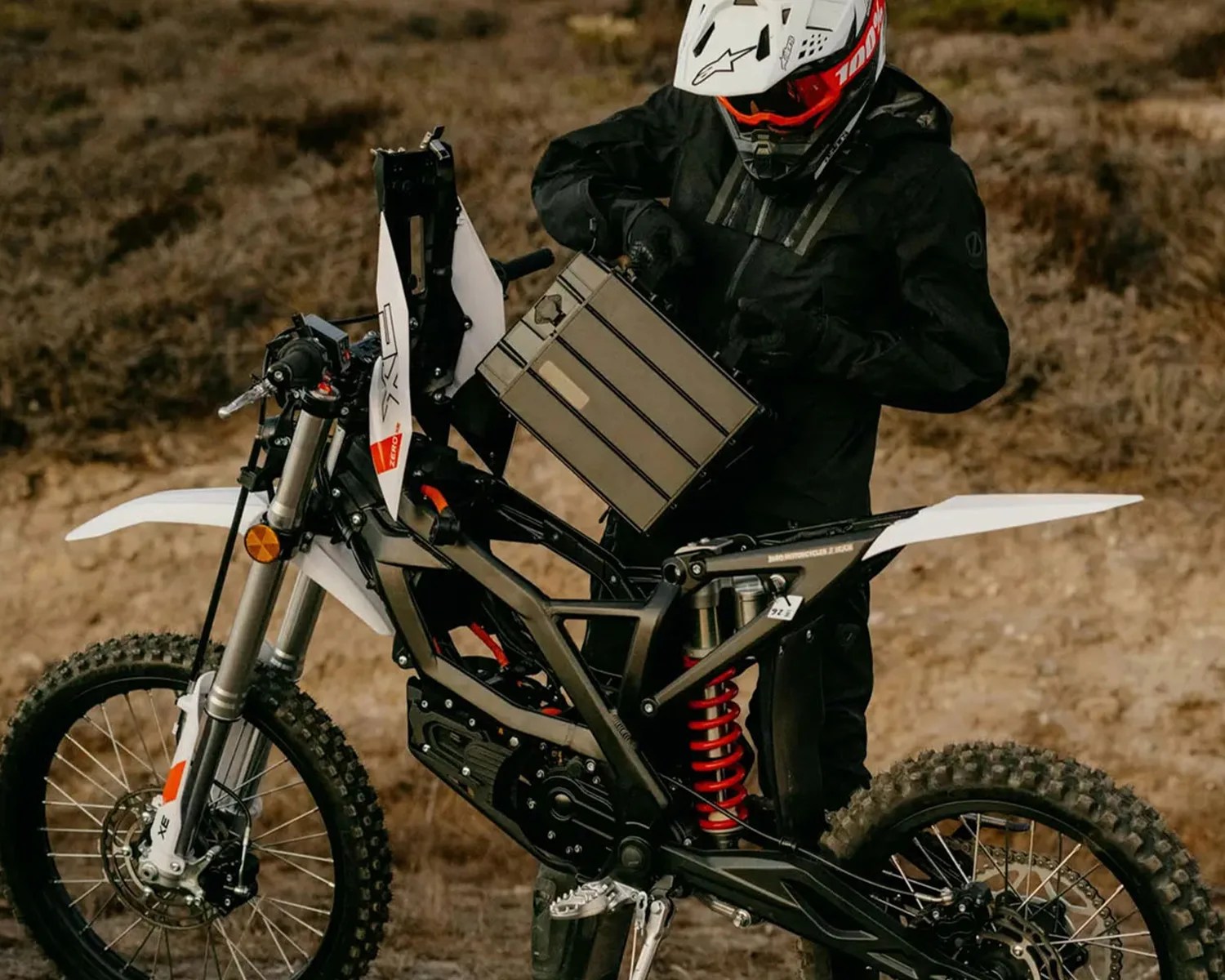 Person in black gear and white helmet holding a large battery above the frame of a black and white electric dirt bike.