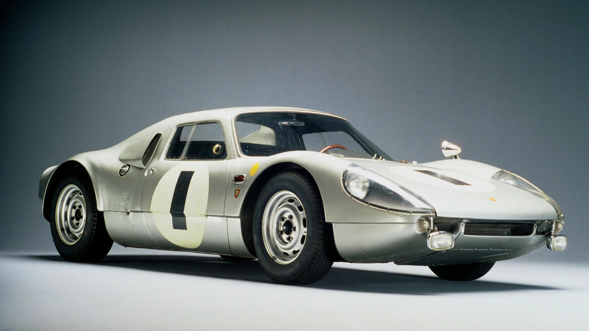 Silver vintage Porsche race car with number 0 on the door and round headlights on a gray background.