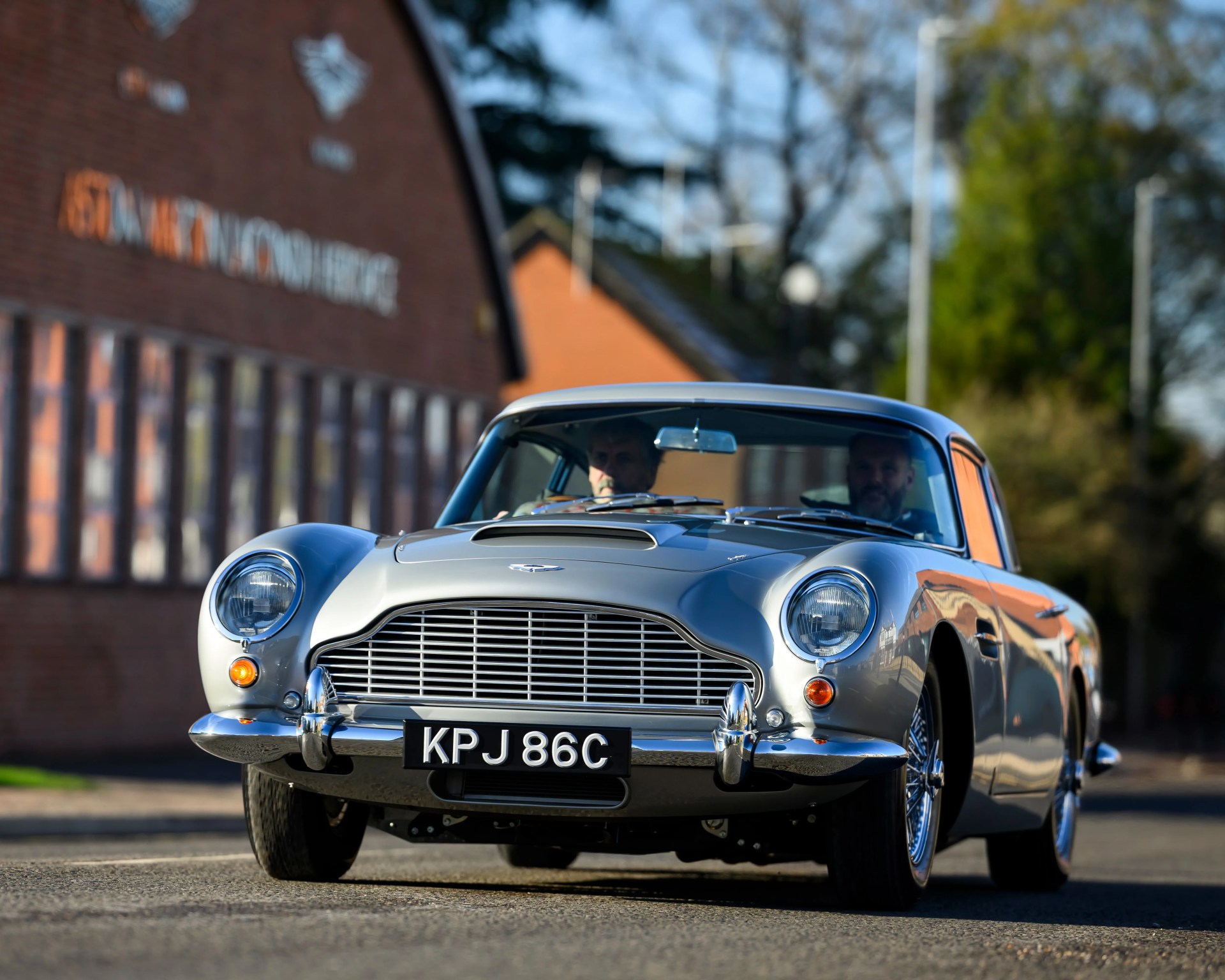 Silver classic Aston Martin car with chrome grille and license plate KPJ 86C on a street.