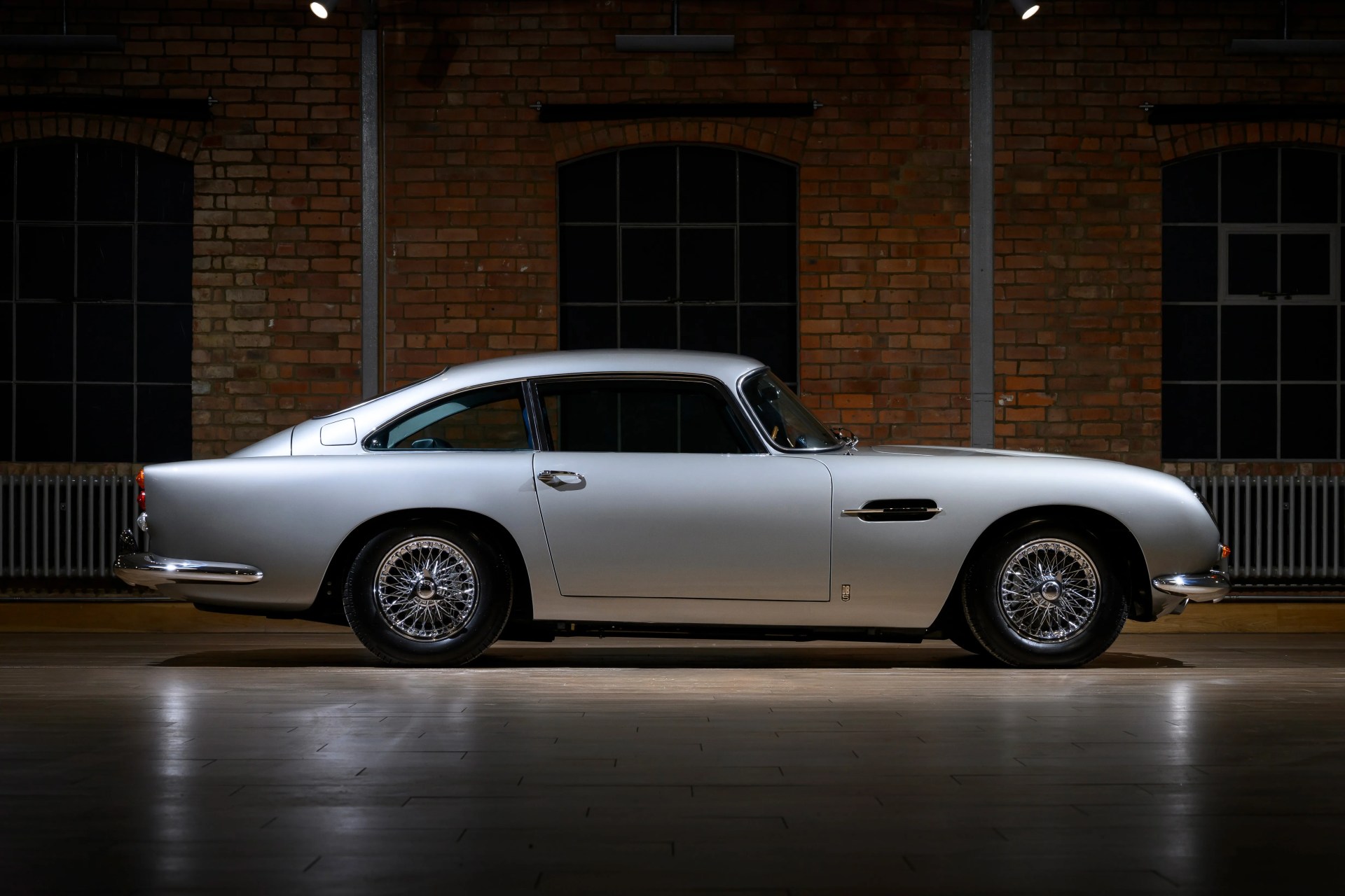 Silver classic Aston Martin DB5 coupe with wire-spoke wheels in an industrial-style room with brick walls.