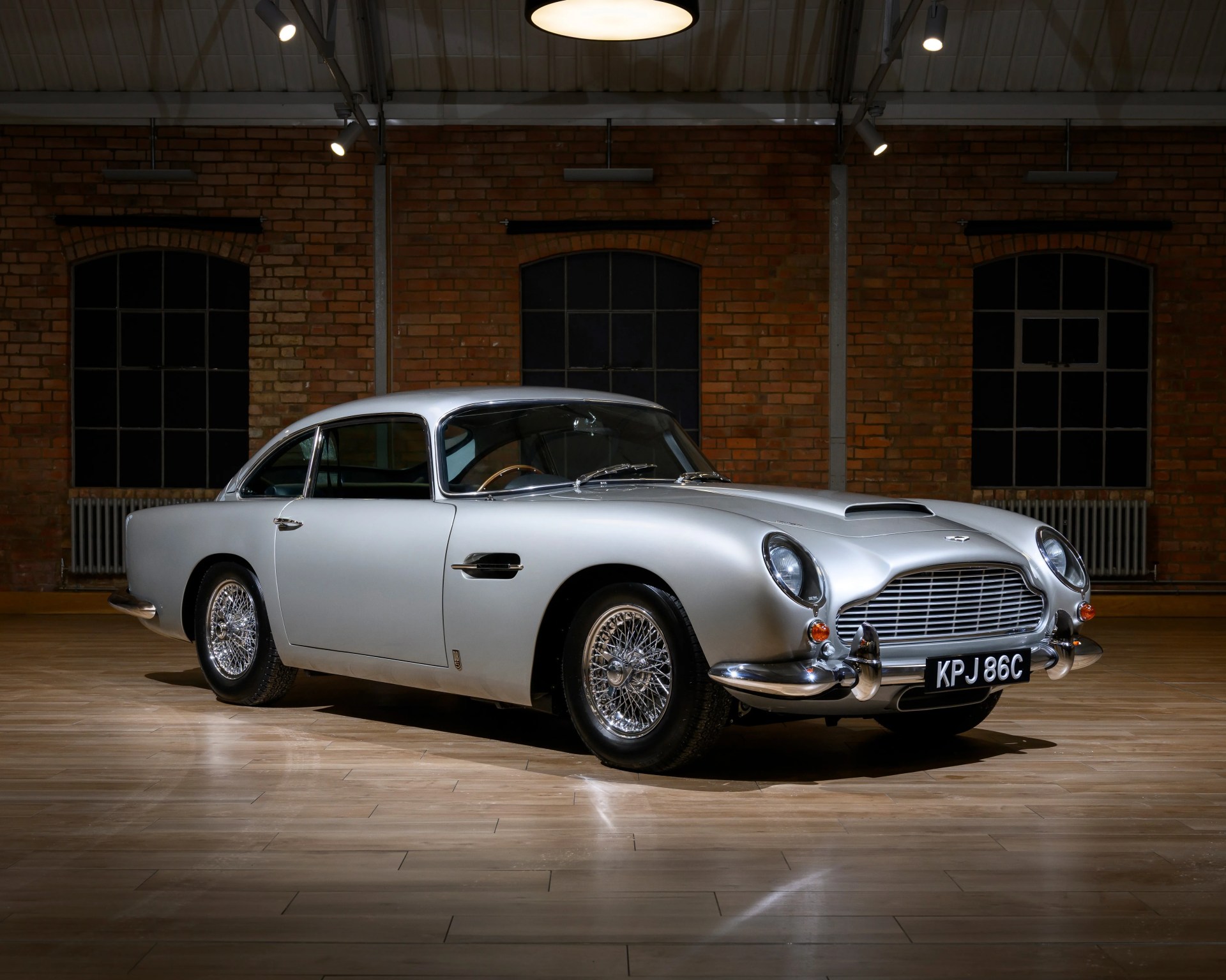 Silver classic Aston Martin DB5 coupe with wire-spoke wheels in an indoor setting with brick walls.