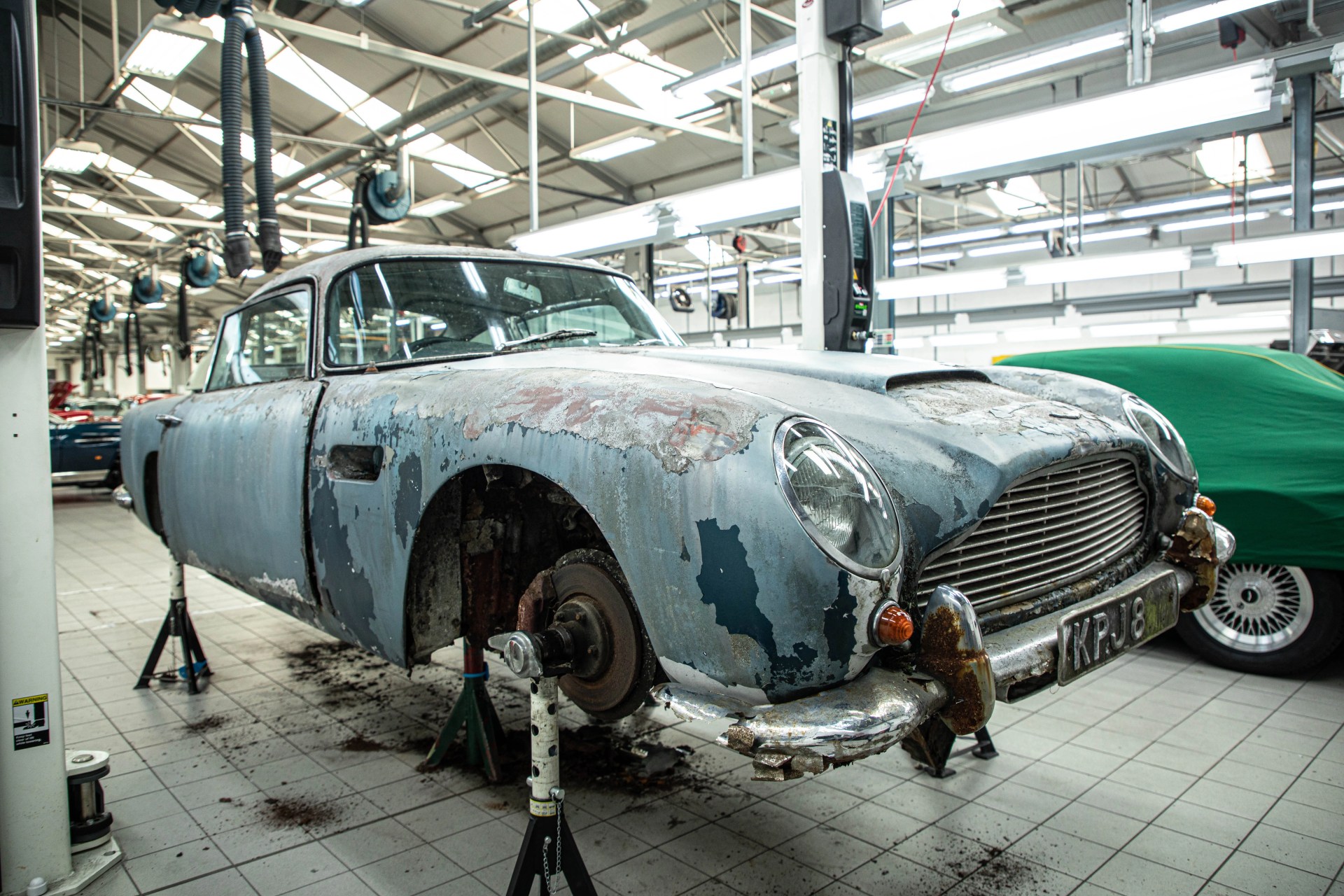 Rusty and paint-peeling classic car without wheels on stands inside a bright garage.