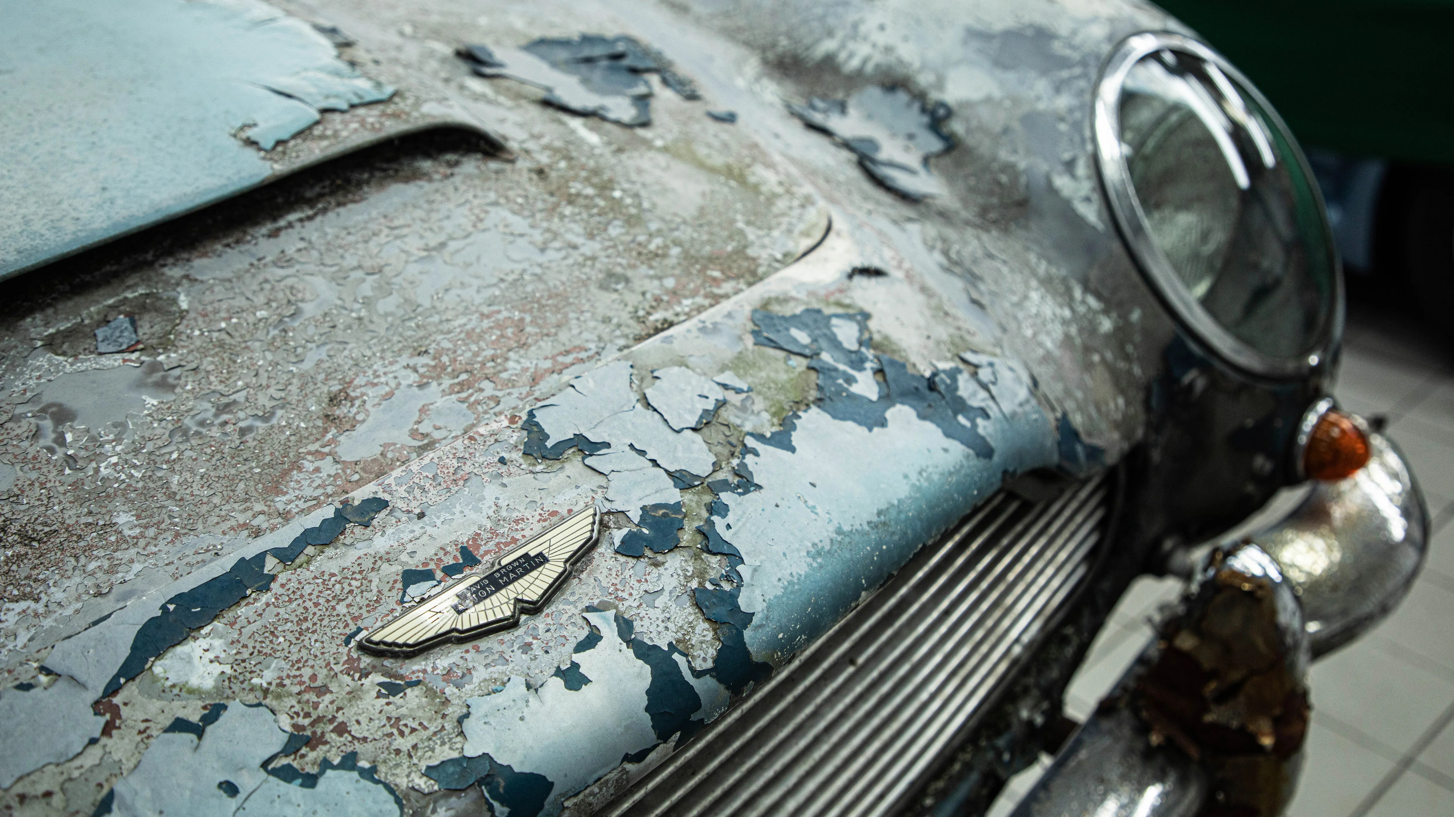 Close-up of a weathered Aston Martin car hood with peeling paint and a rusty front bumper.