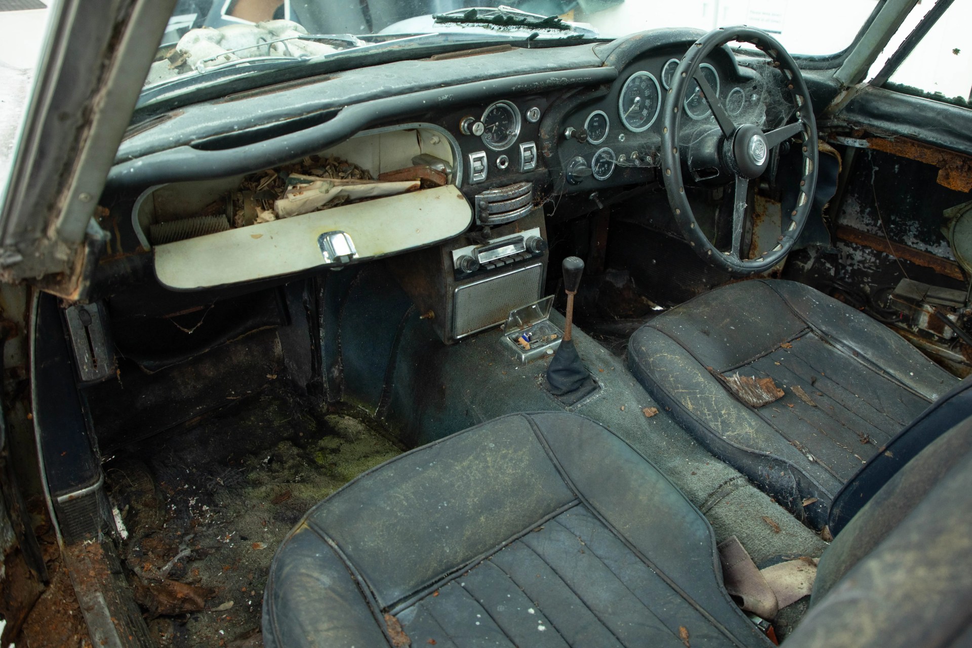Interior of an old, dusty car with worn black seats, an open glove compartment filled with debris, and a steering wheel covered in cobwebs.