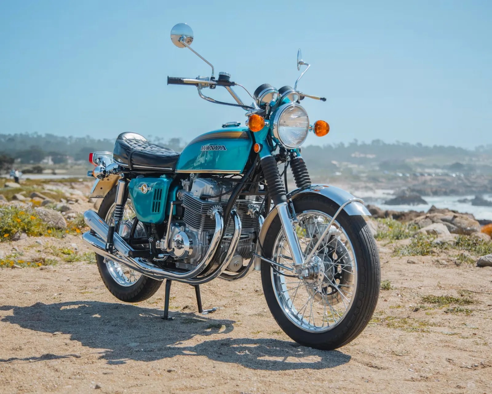 Vintage teal Honda motorcycle with chrome details parked on sandy ground near rocky coastline.