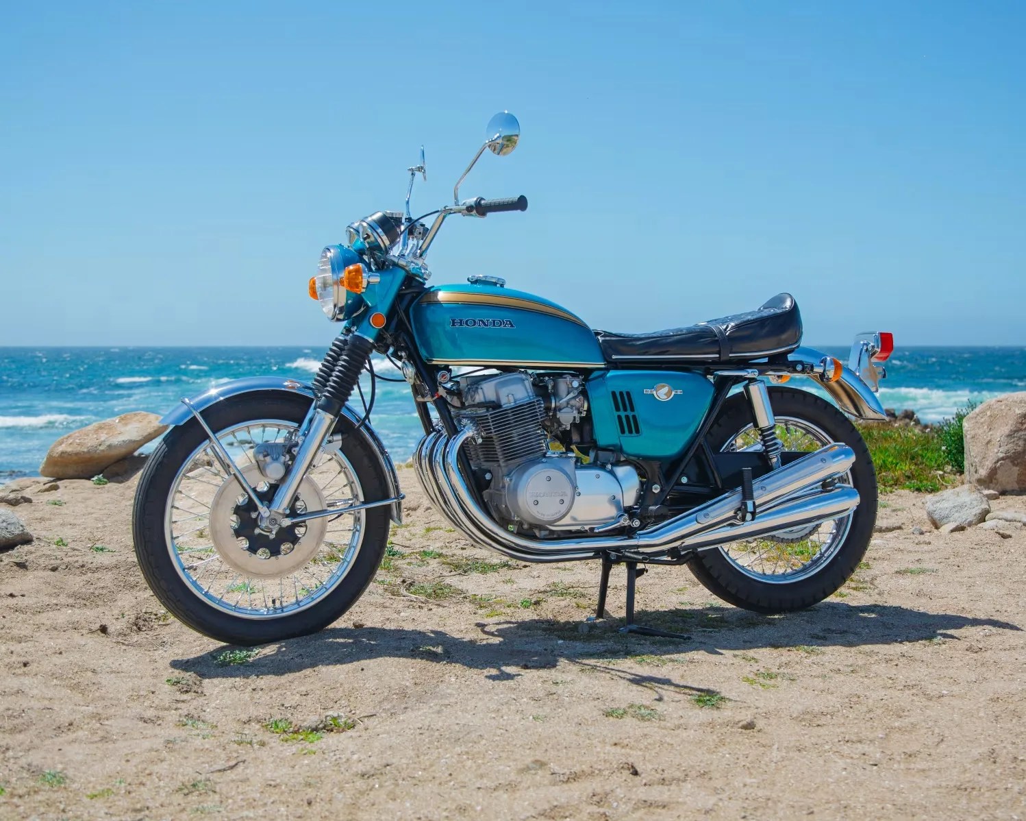 Teal vintage Honda motorcycle with chrome exhaust pipes parked on sandy beach near ocean.