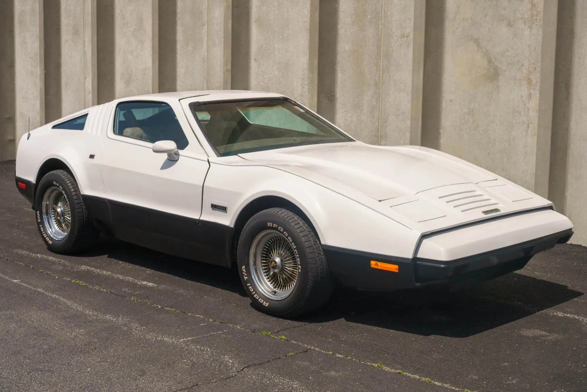 White and black 1970s sports car with wire-spoke wheels parked on asphalt near a concrete wall.