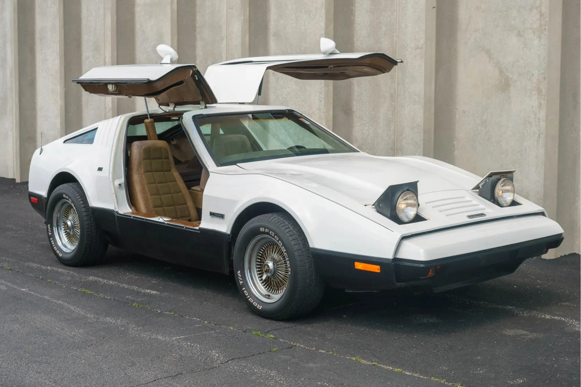 White sports car with gullwing doors open and pop-up headlights raised, parked on asphalt near a concrete wall.