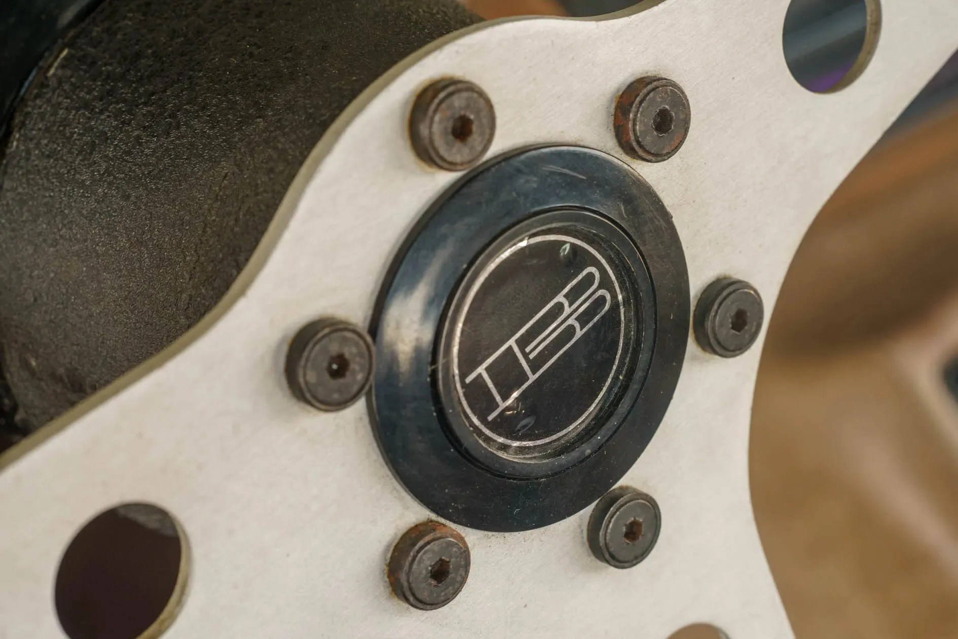 Close-up of a white metal wheel hub with six rusty bolts and a black center cap featuring a stylized "HB" logo.