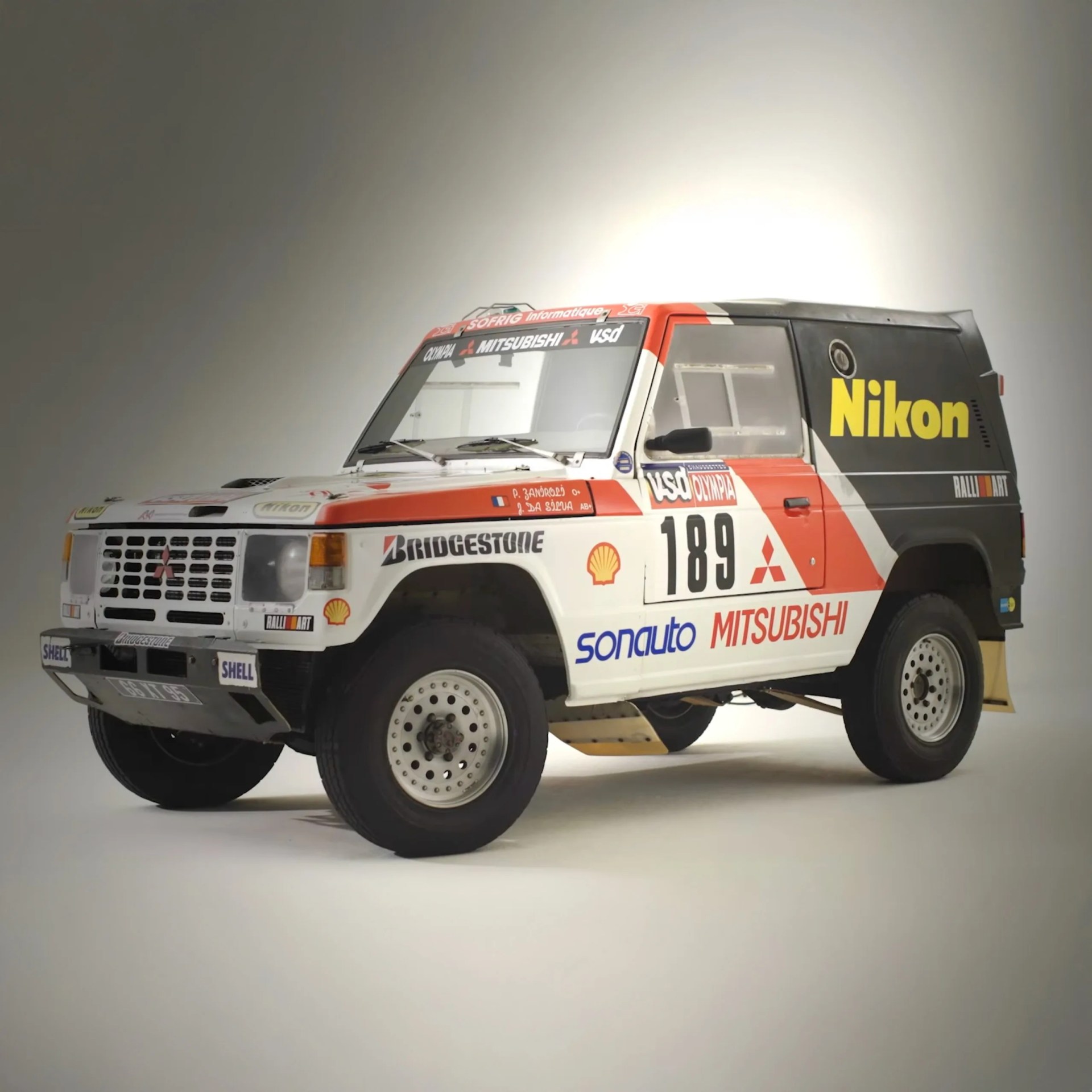 White and red Mitsubishi rally car with Nikon and Bridgestone logos, number 189, on a plain background.