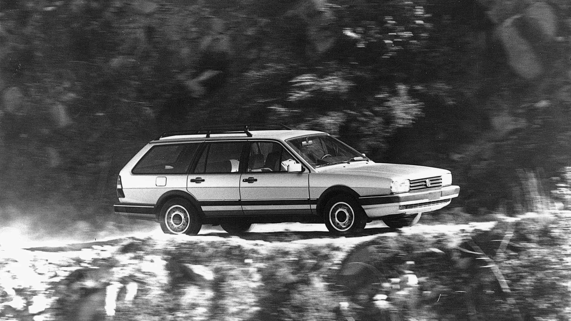 Black and white photo of a vintage station wagon driving on a dirt road with blurred forest background.