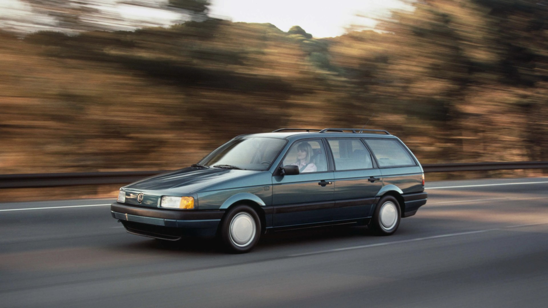 Dark green Volkswagen station wagon driving on a highway with blurred autumn trees in the background.