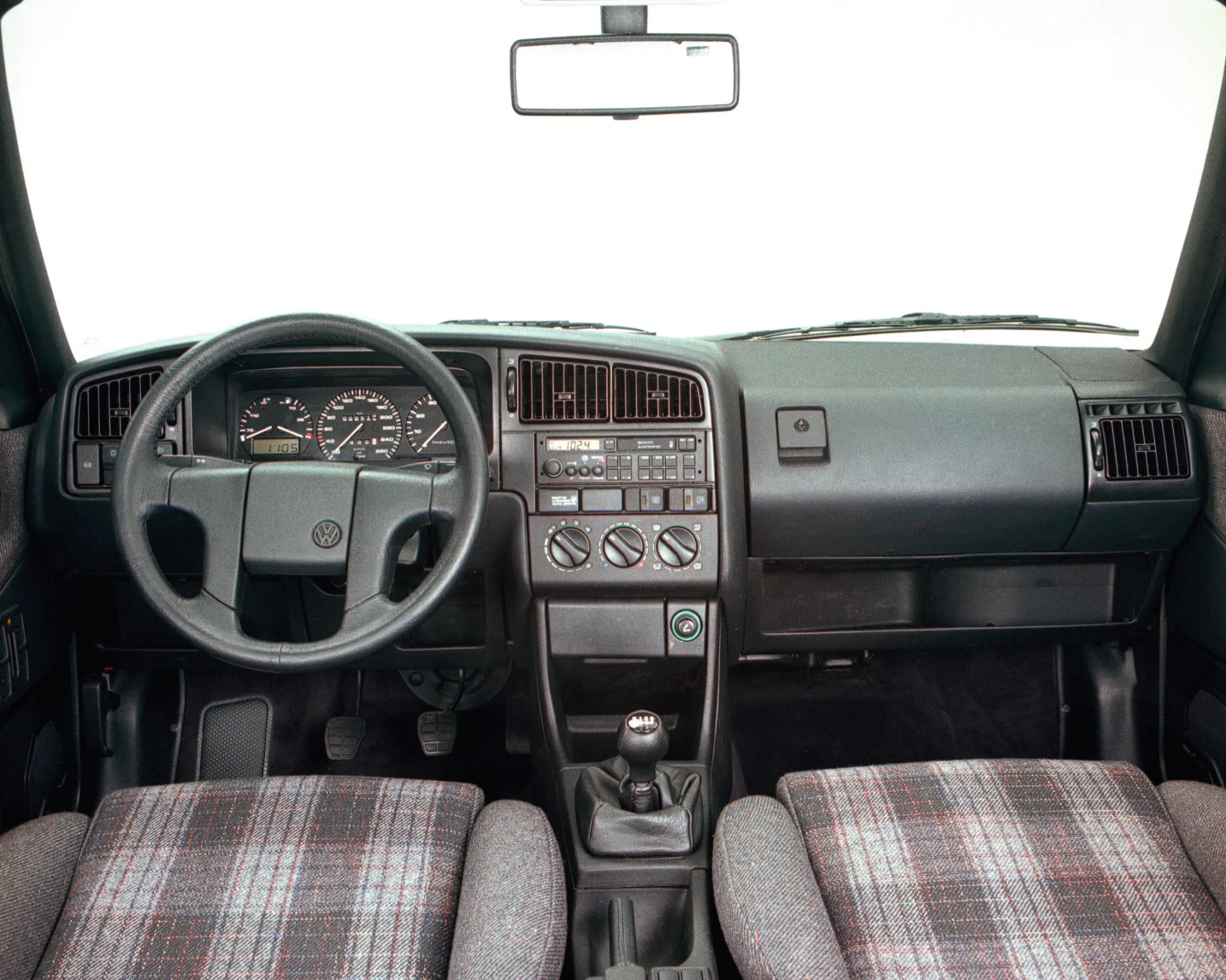 Dashboard and steering wheel of a Volkswagen car with plaid fabric seats and manual gear shift.