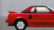 Red two-door sports car with rear spoiler and white wheels parked against a gray wall.