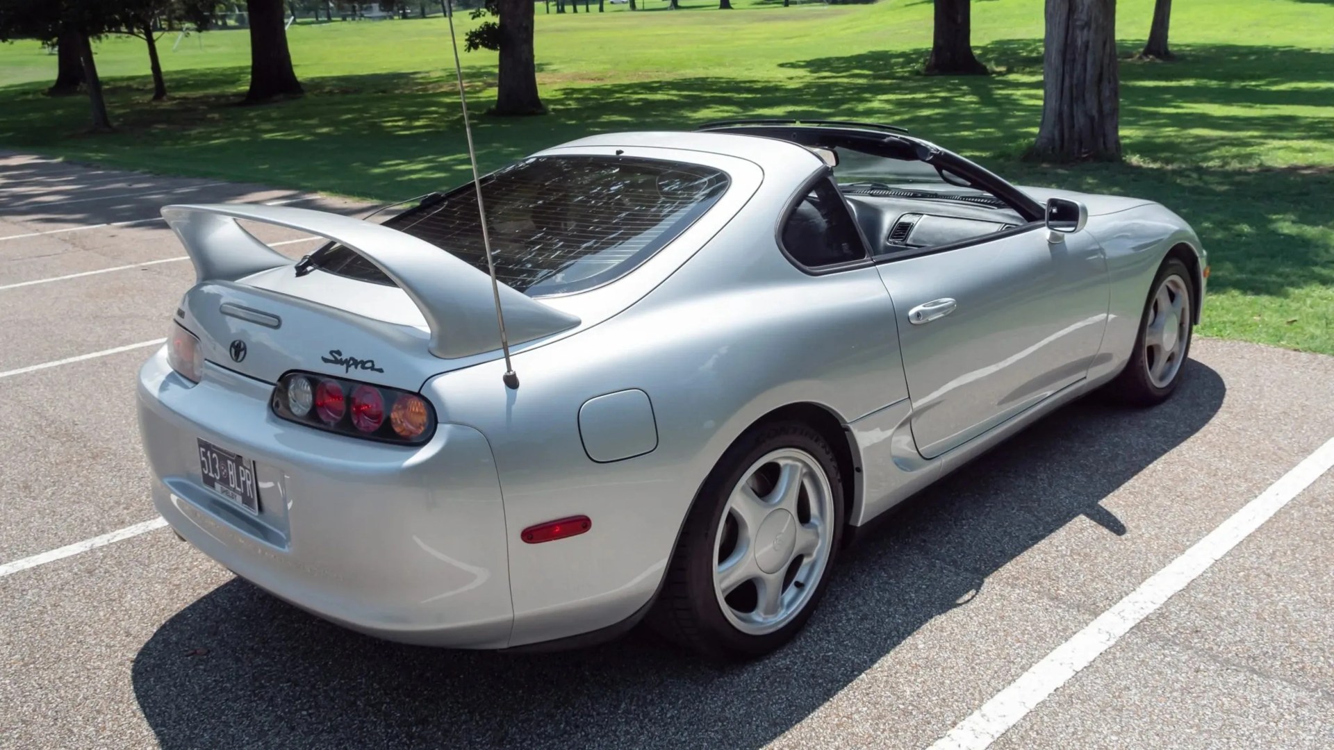 Silver Toyota Supra sports car with rear spoiler parked in a sunlit outdoor lot near grass and trees.