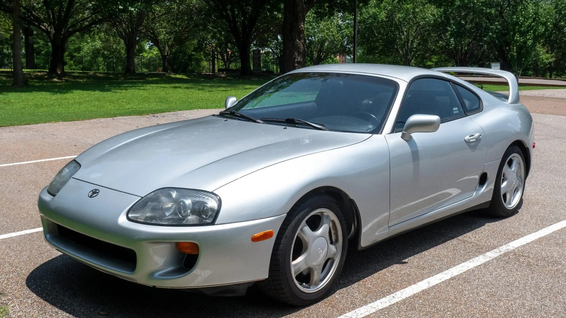 Silver Toyota Supra sports car parked in a lot with trees in the background.
