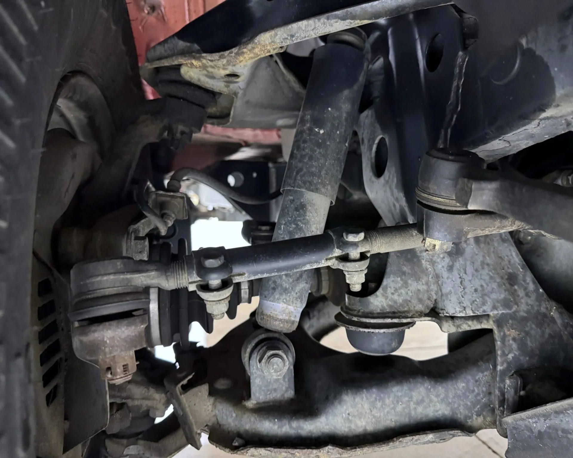 Close-up of a vehicle's suspension system showing a shock absorber, control arms, and tie rod ends with visible bolts and dust.