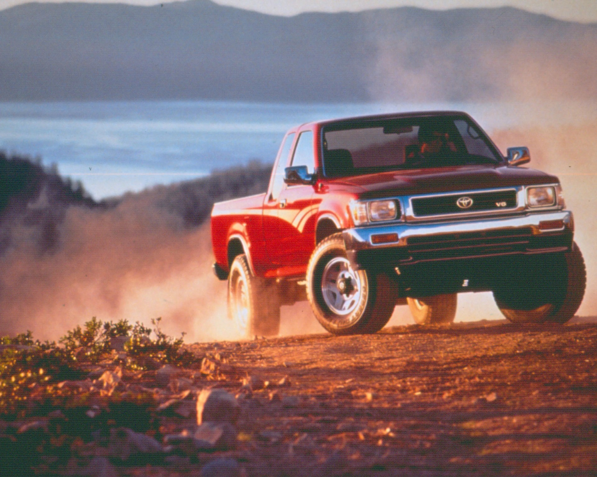 Red Toyota pickup truck kicking up dust on a dirt road with mountains and a lake in the background.