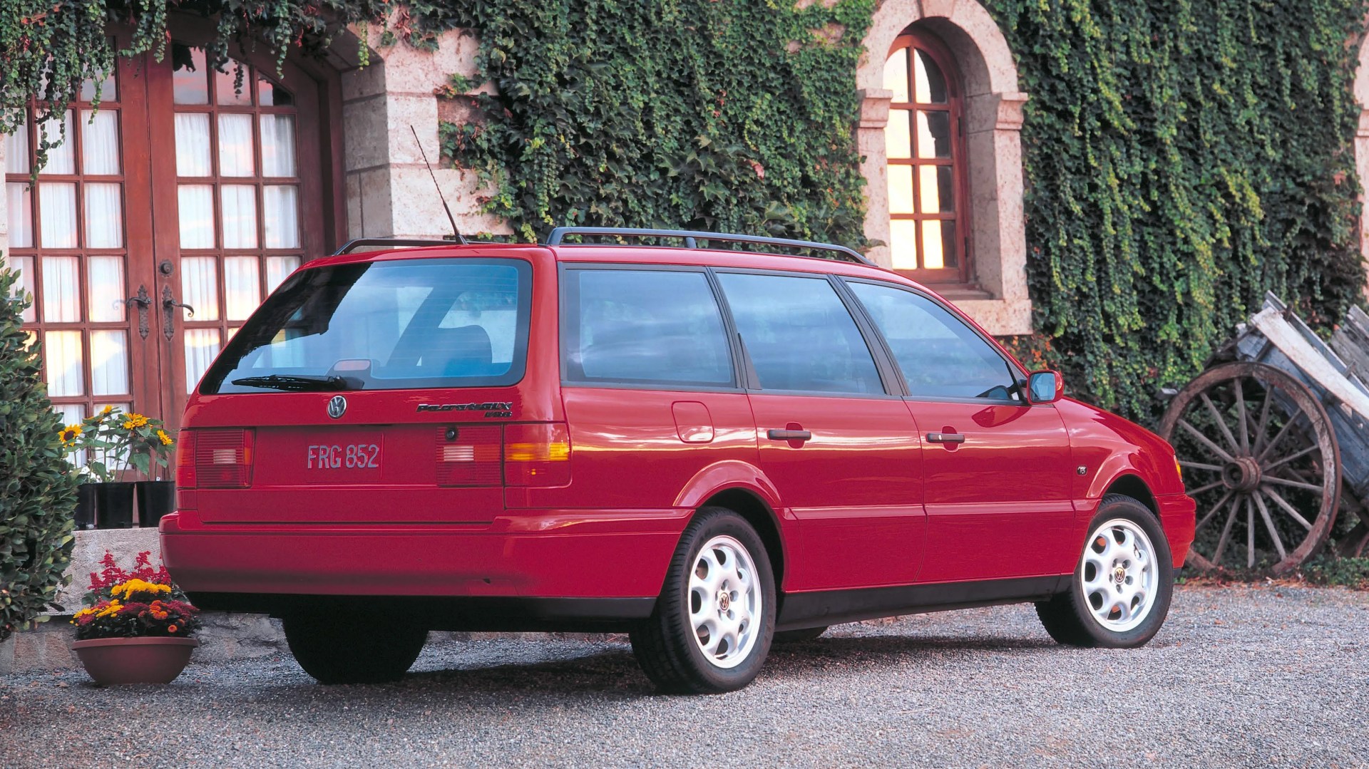 Red Volkswagen Passat station wagon parked on gravel in front of a stone building with ivy and wooden cart wheels.