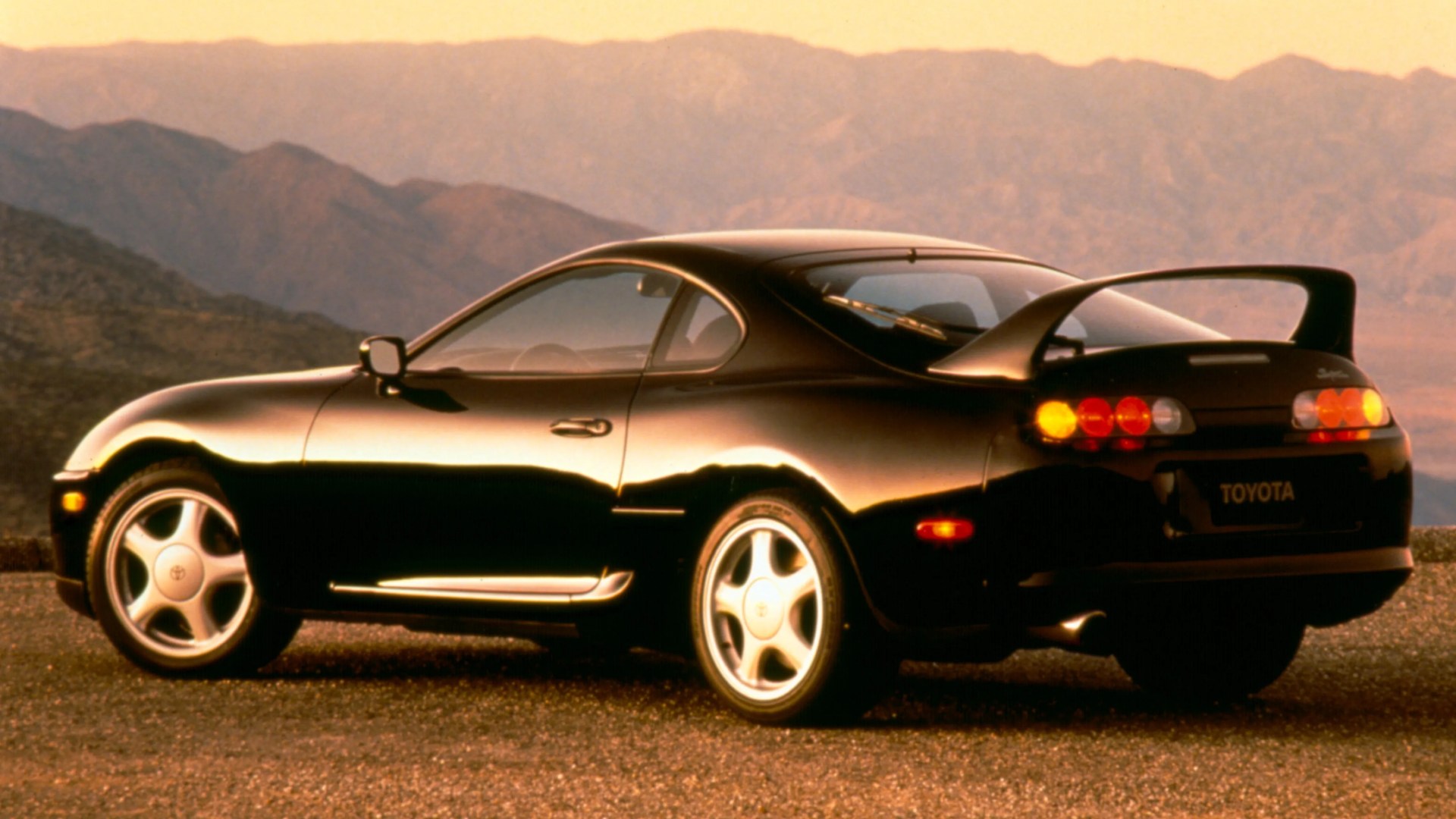 Black Toyota Supra sports car with rear spoiler parked on a gravel surface with mountains in the background.