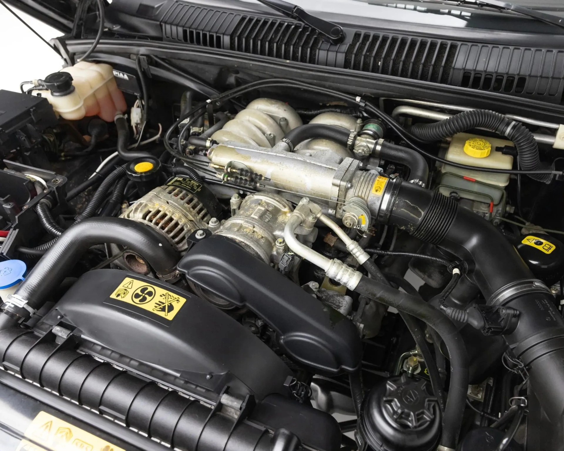 Car engine compartment showing various hoses, pipes, and components with yellow warning labels.