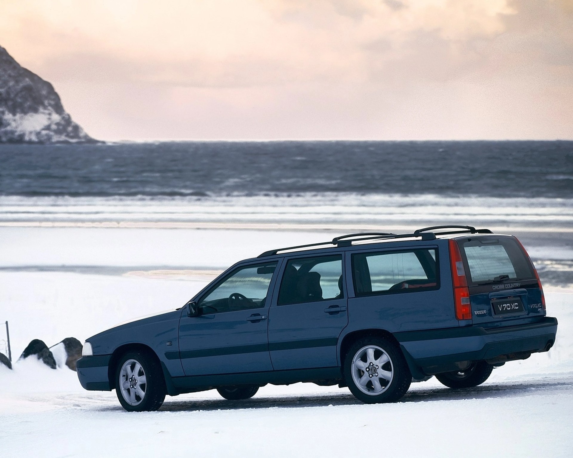 Blue Volvo V70 XC Cross Country station wagon parked on snowy ground near a body of water with rocky hills in the background.