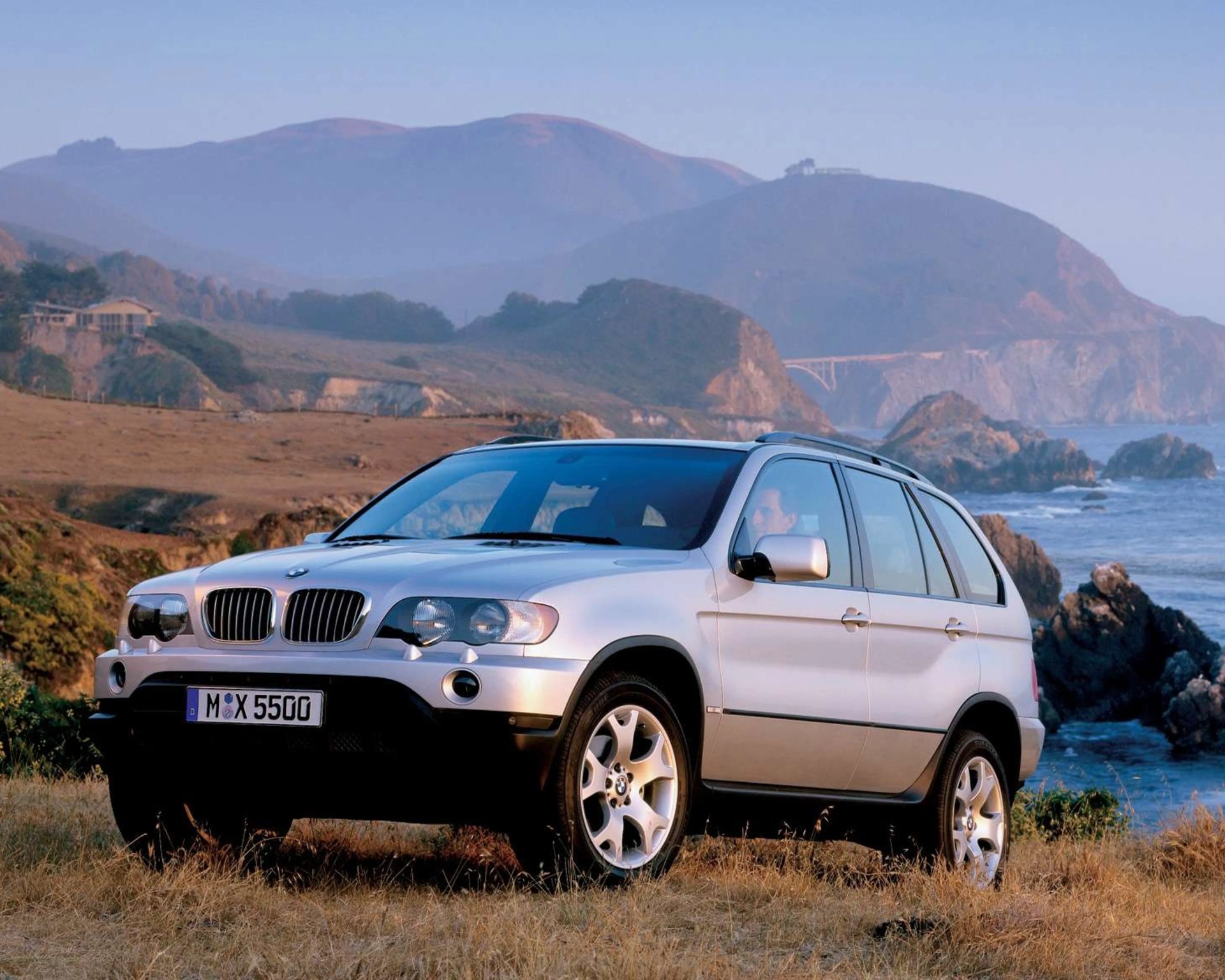 Silver BMW X5 SUV parked on dry grass with rocky coastline and hills in the background.