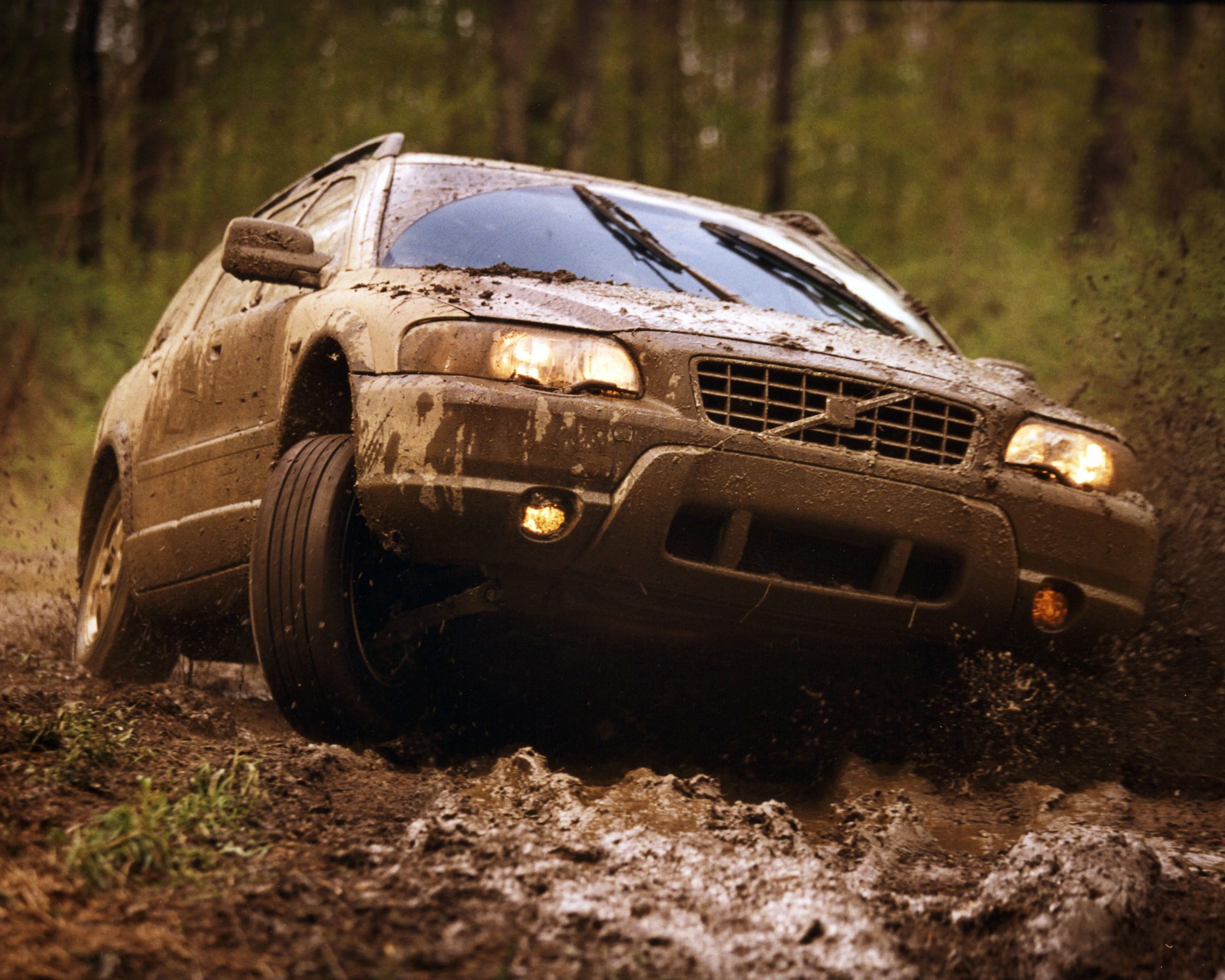 Mud-covered beige Volvo station wagon driving through a muddy forest trail with headlights on.