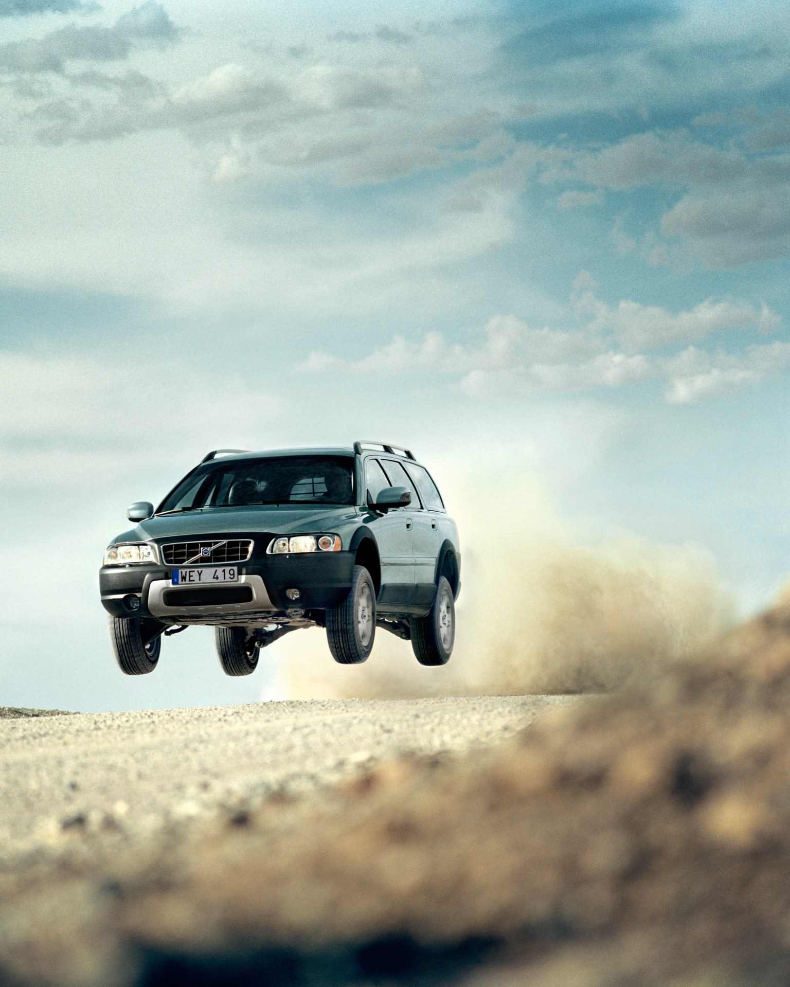 Dark green Volvo SUV airborne on a dirt road with dust trailing behind under a cloudy sky.