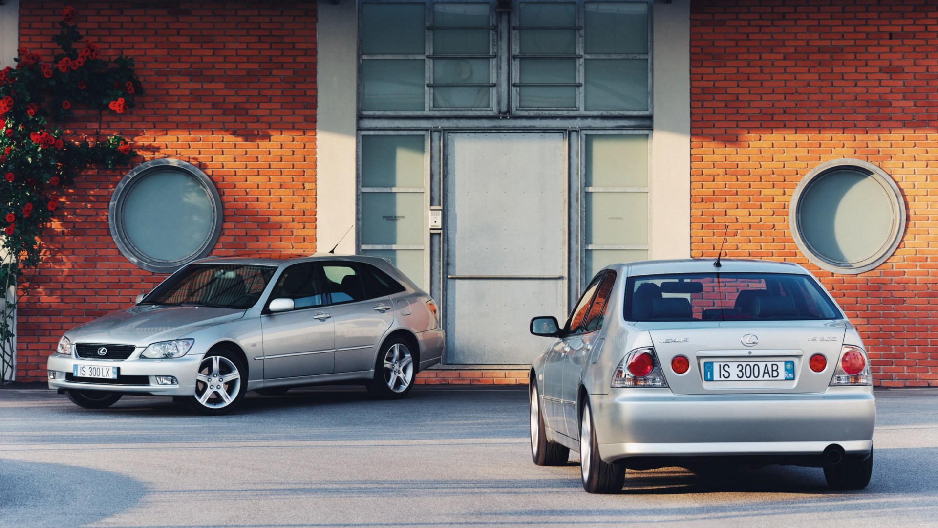 Two silver Lexus IS 300 cars parked in front of a brick building with circular windows and red flowers on the left side.