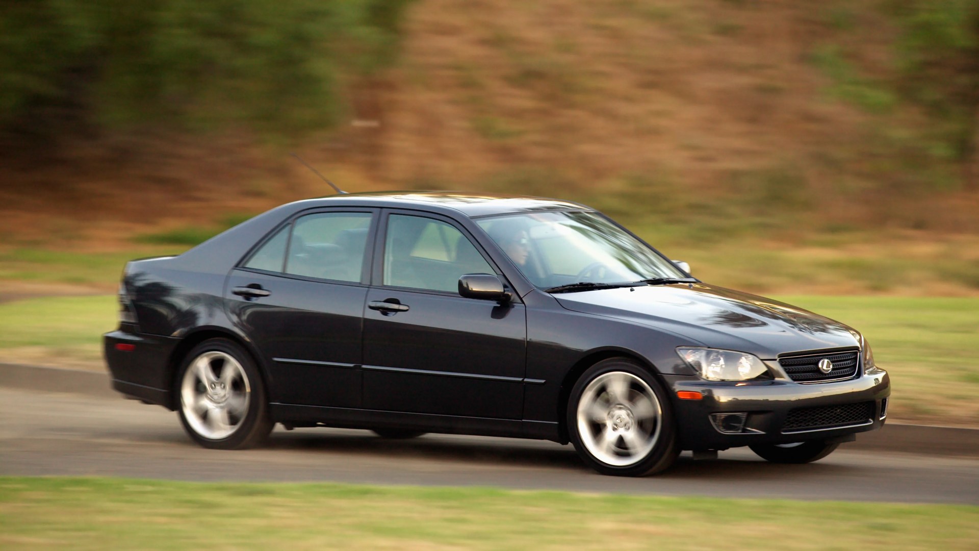 Black Lexus sedan with silver alloy wheels driving on a paved road with blurred background.