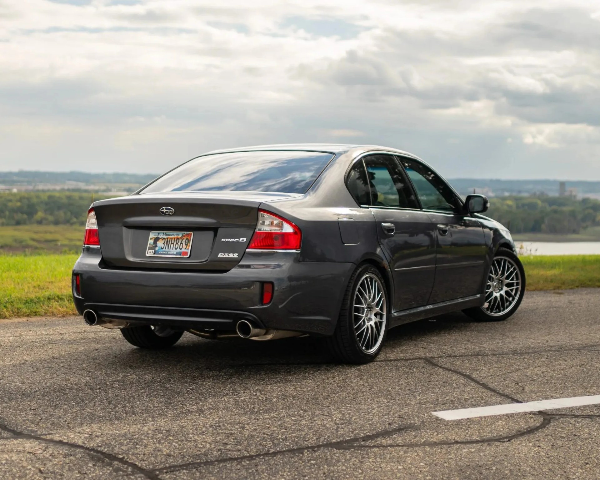 Dark gray Subaru sedan with aftermarket multi-spoke wheels parked on a road with grass and cloudy sky in the background.