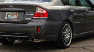 Rear side view of a dark gray Subaru Spec B sedan with Minnesota license plate and multi-spoke alloy wheels.