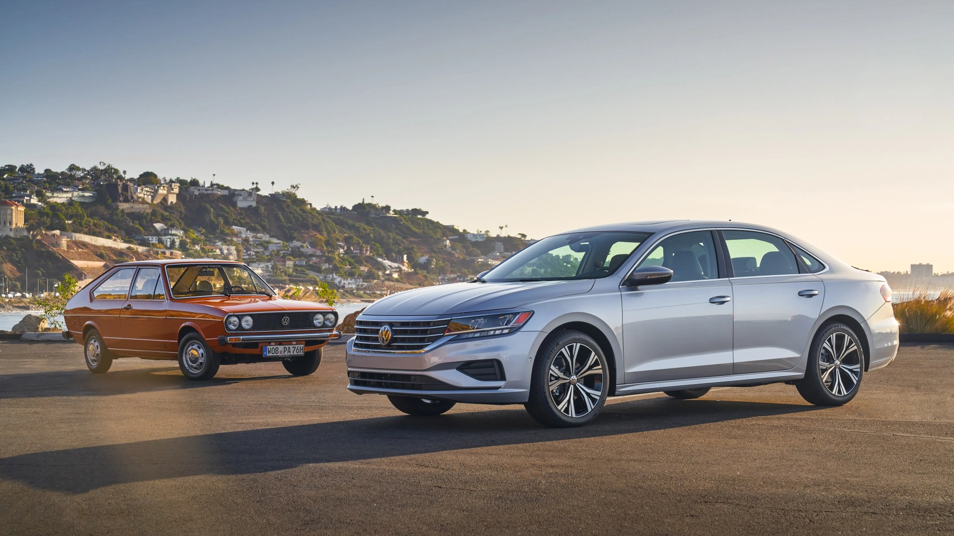 Silver modern Volkswagen sedan parked next to a vintage orange Volkswagen hatchback on a paved area with hills in the background.