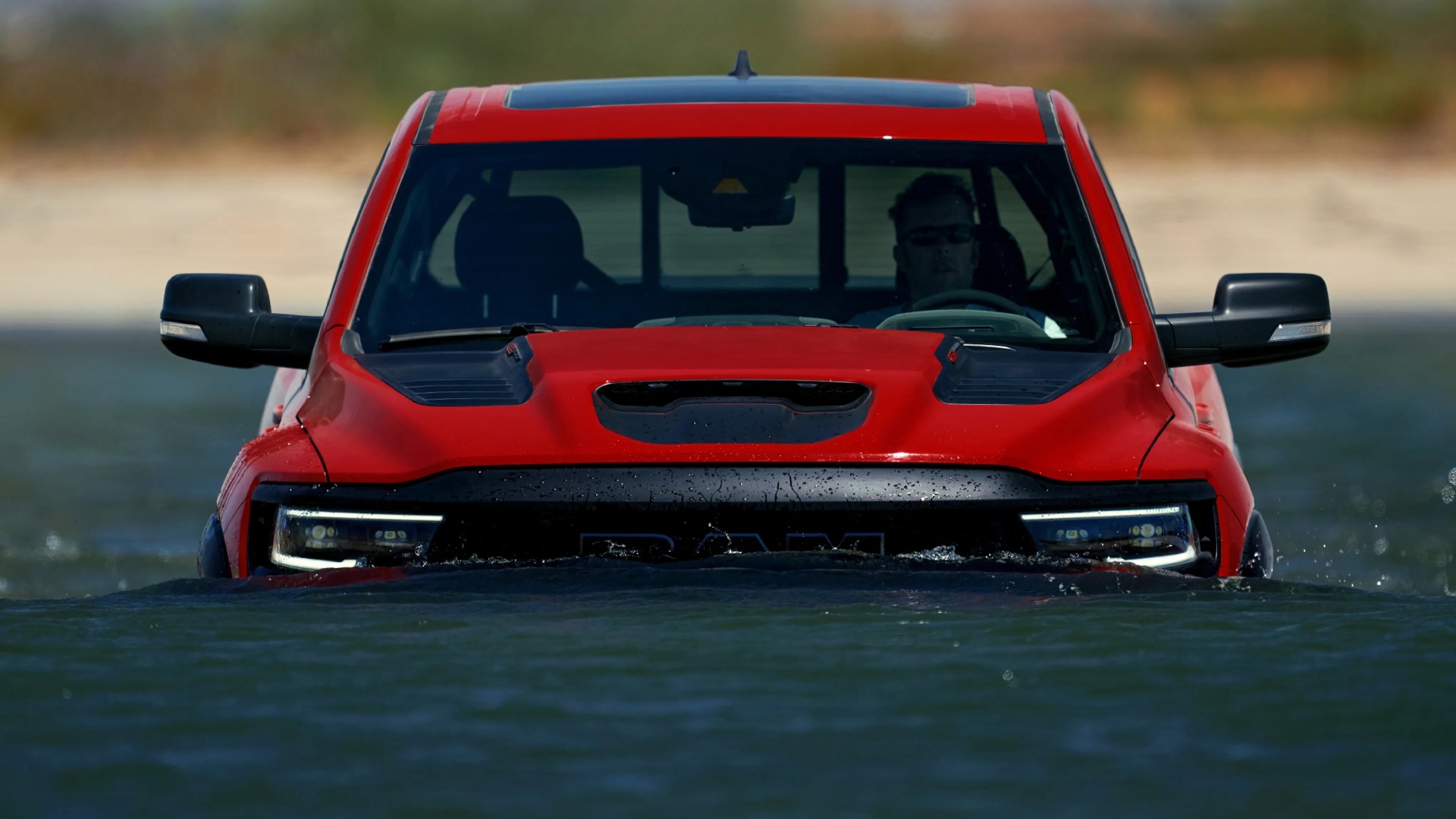 Red pickup truck partially submerged in water with a person visible inside wearing sunglasses.