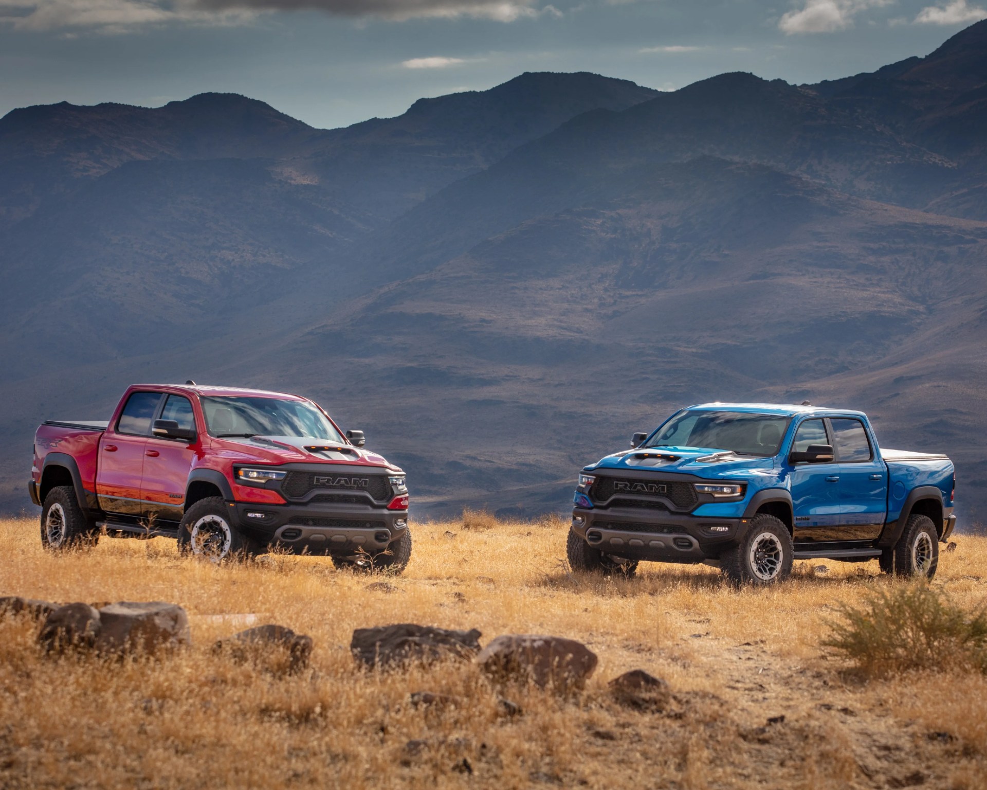 Two RAM pickup trucks, one red and one blue, parked on dry grass with mountains in the background.